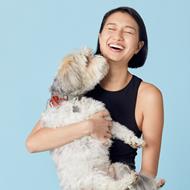 Womam sitting on a stool holding her dog. The dog is licking her face and she is laughing