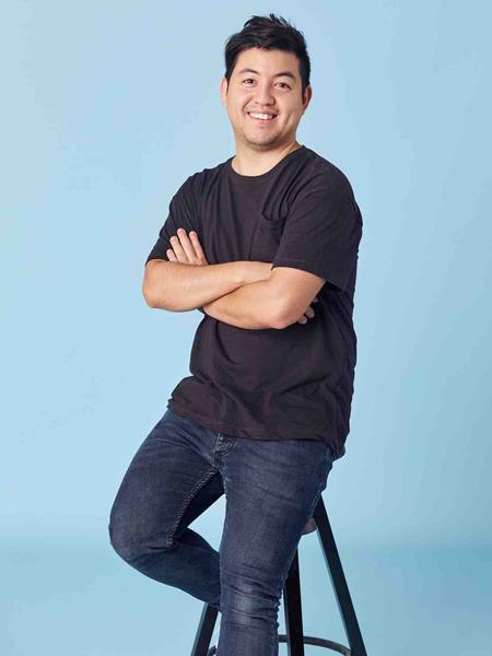 Man in a black t-shirt with his arms folded smiling to the camera and sitting on a stool against a blue background