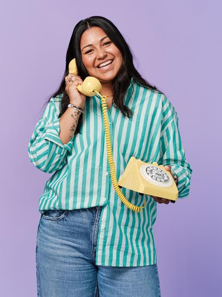 Medium skinned woman wearing green and white striped shirt and jeans, holding an old fashioned yellow telephone.