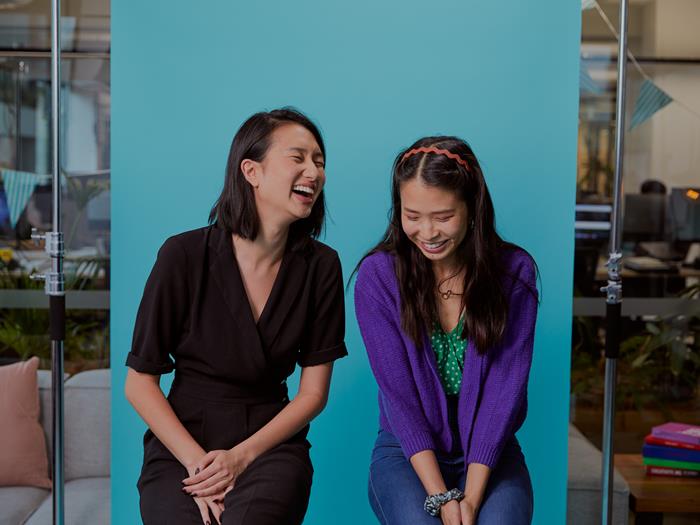 Two women sitting on stools against a blue background laughing and smiling down towards the floor