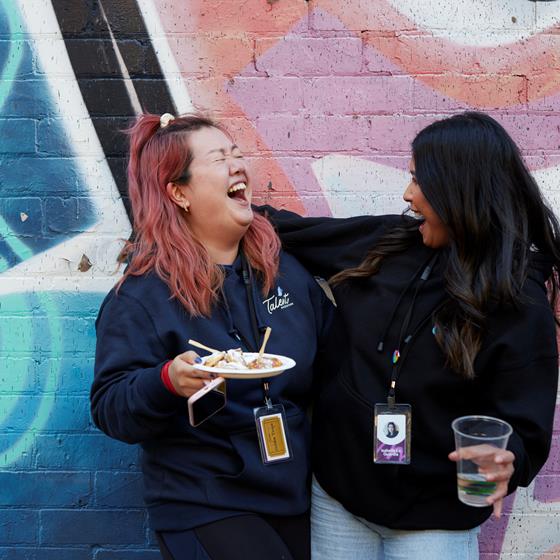 Two women smiling and laughing at each other holding a plate of food and a cup of drink, standing in front of a coloured brick wall
