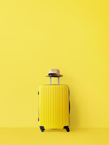 A yellow suitcase against a yellow background with a hat on the suitcase carry handle