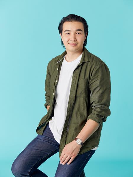 Light skinned man in jeans, white t shirt and olive green shirt, sitting on a stool.