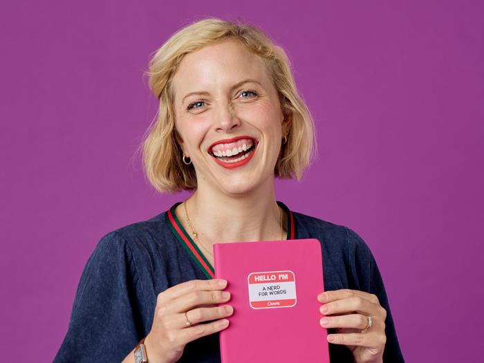 Light skinned woman in blue dress holds a pink notebook with a sticker on it saying, 'Hello I'm a nerd for words'.
