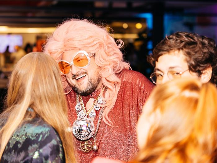 Light skinned man in drag with a disco ball necklace and wig talking to a group of people.