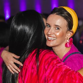 Light skinned woman with bright head band, earrings and top, hugging person with long dark hair.