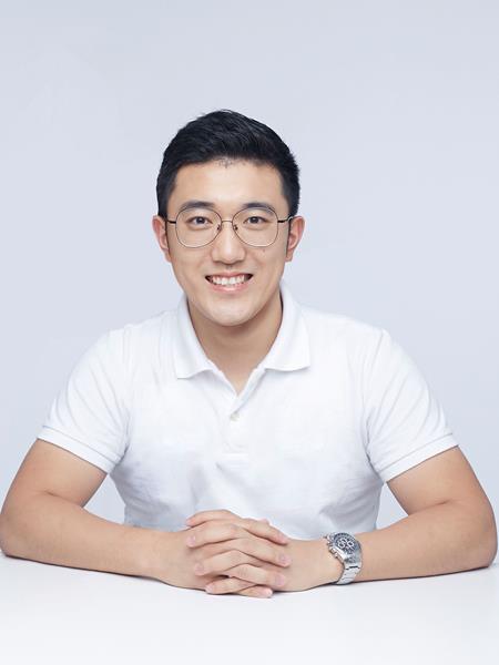 Man sitting at a white desk with linked hands and elbows resting on the desk. Wearing a white shirt and glasses