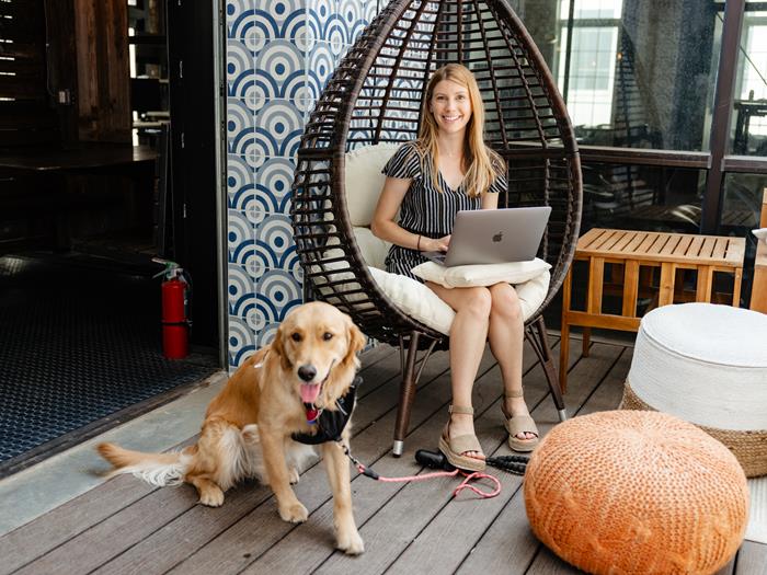 Women sitting in a hanging chair working on her laptop and smiling to the camera. Her dog on a leash sitting next to her.