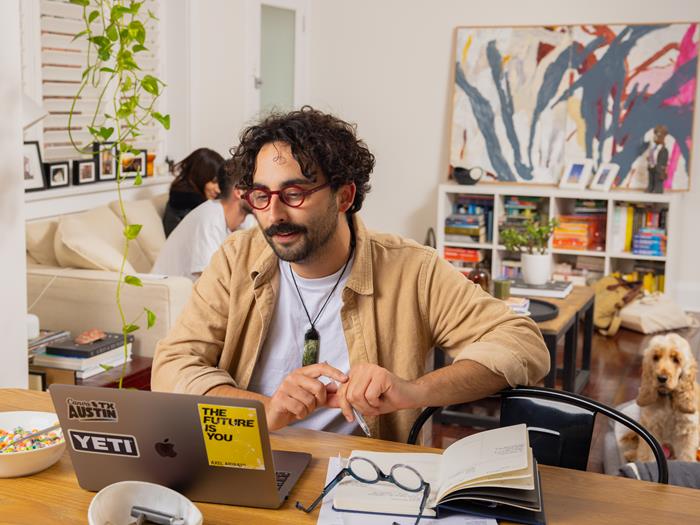 Man at his kitchen table working on his laptop with a bowl of cereal next to him and his dog sitting on the floor in the background