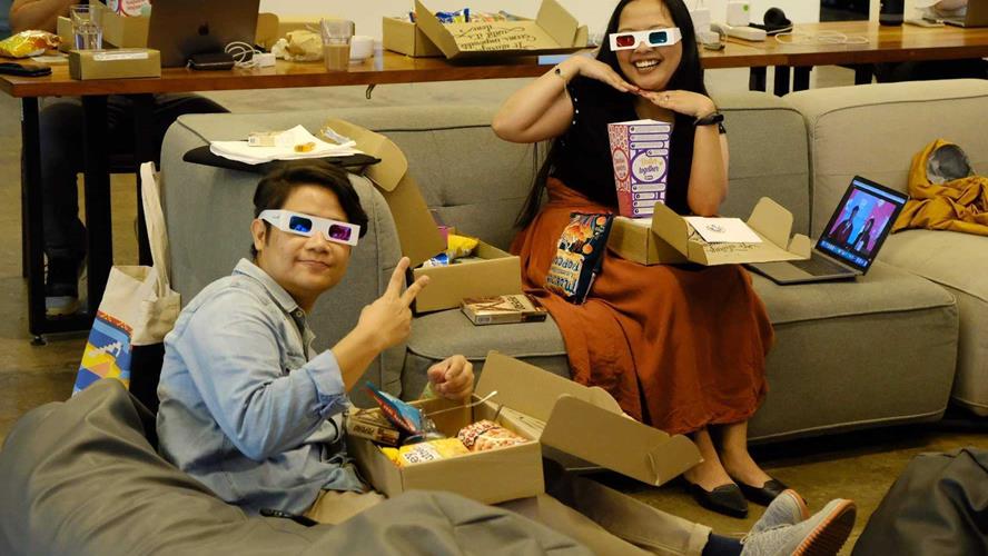 Two Canva colleagues sitting on a couch and a bean bag in the office eating popcorn and wearing 3D glasses.