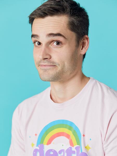Man against a blue background with a white t-shirt with a rainbow on it smiling to the camera