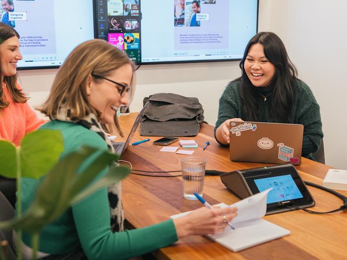 Three women sitting in a meeting room at a desk on their laptops smiling.