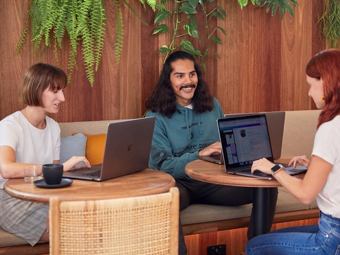 Woman in white tee shirt sits at cafe table working on laptop. At the table next to her a man in a green hoodie and a white tee shirt collaborate on laptops.