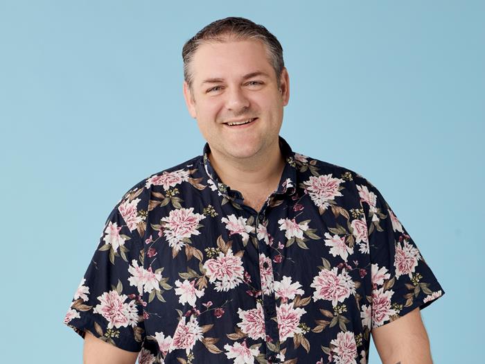 Light skinned man in floral shirt and black trousers, smiling.