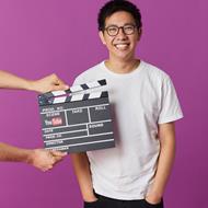 Man in a white t-shirt and glasses facing front on to camera and smiling. A movie action sign held in front of him.