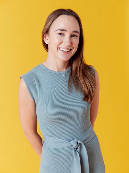 Women with blue dress and long brown hair with hands behind her back smiling at the camera