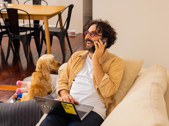 Man on the phone, sitting on his couch with his laptop on his lap. Dog sitting next to him.