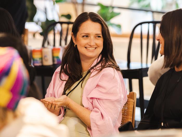 Light skinned woman in a pink shirt sitting down and smiling at another women with her back to the camera.