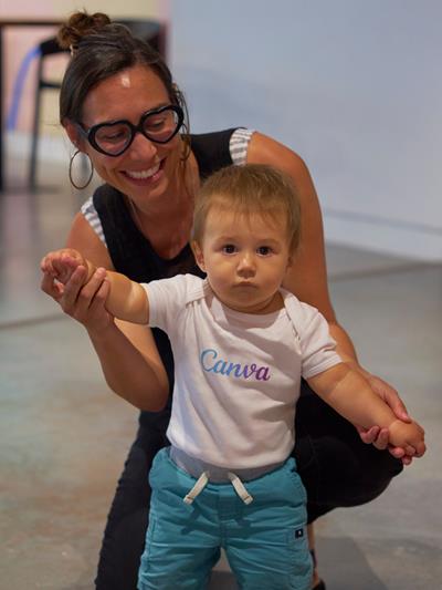 Medium skinned woman kneeling on a concrete floor, holding the hands of a toddler standing in front of her.