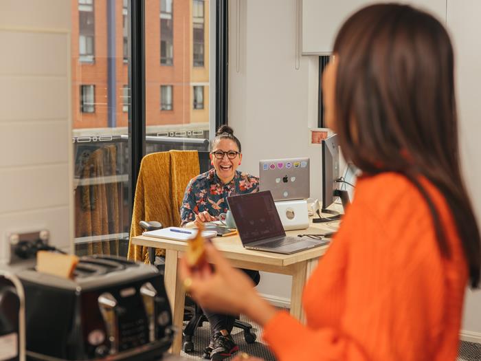 Women in the background sitting at her desk working on her laptop. Facing the camera and laughing to the women in the foreground who is eating toast and wearing an orange jumper