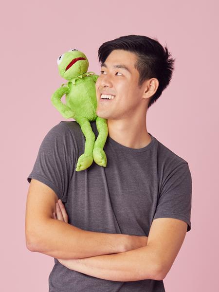 Man in dark grey shirt with arms crossed and frog plush toy on left shoulder. Man smiling at frog.