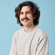 Man with curly brown hair and moustache sitting and smiling in front of a blue background.