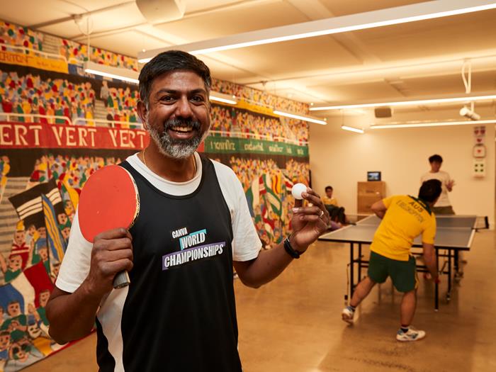 Man holding ping pong ball and bat and two men playing ping pong in the background