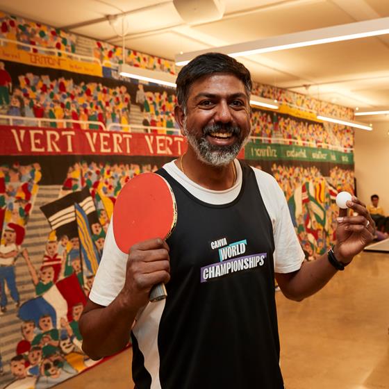Man holding ping pong ball and bat and two men playing ping pong in the background