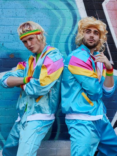 Two men dressed in bright tracksuits and blonde wigs standing in front of a colourful wall