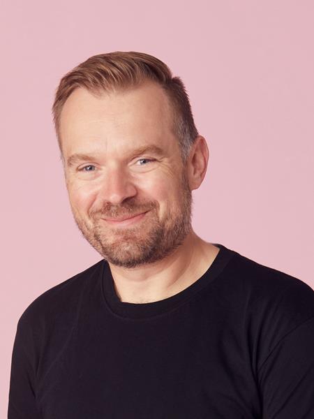 Man in a black t-shirt against a pink background smiling to the camera