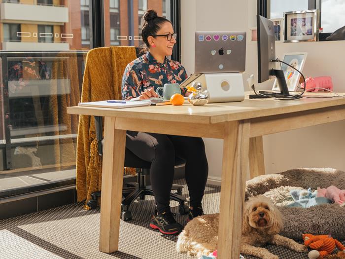 Women sitting at her desk working on her laptop. Dog sitting at the base of her feet.