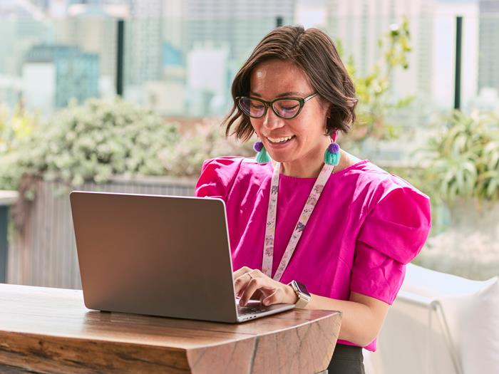 A light skinned woman in a bright pink top working at a laptop on a wooden bench outside.