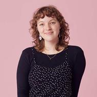 Women with curly brown hair wearing a black long sleeved top with a spotted dress over the top smiling at the camera
