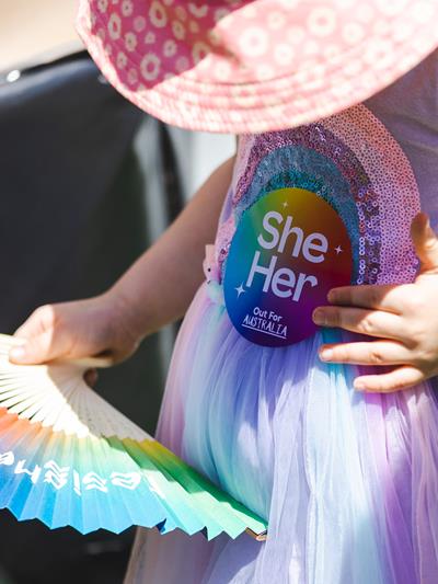 Young girl holding a colourful fan looking down from the camera under hat. Wearing a pride badge that says 'she/her'