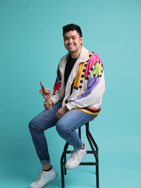 A light skinned man in jeans, a black t shirt and colourful jacket, sitting on a stool smiling and holding a prop.