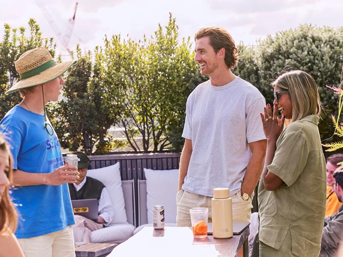 Two light skinned men and a dark skinned woman talking and laughing outside around a table.