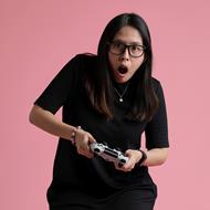Medium skinned woman in a black dress sitting on a stool with a game console controller in her hands.