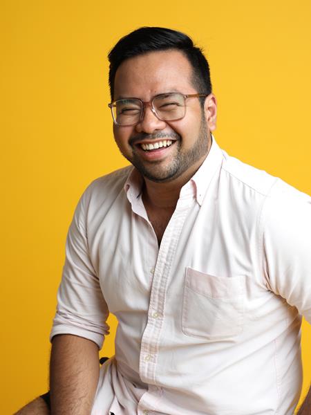 Man with white shirt and glasses sitting on a stool, smiling at the camera. Yellow background