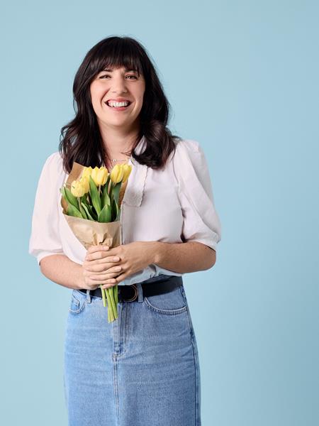 Light skinned woman in white top and denim skirt holding a bunch of yellow tulips.